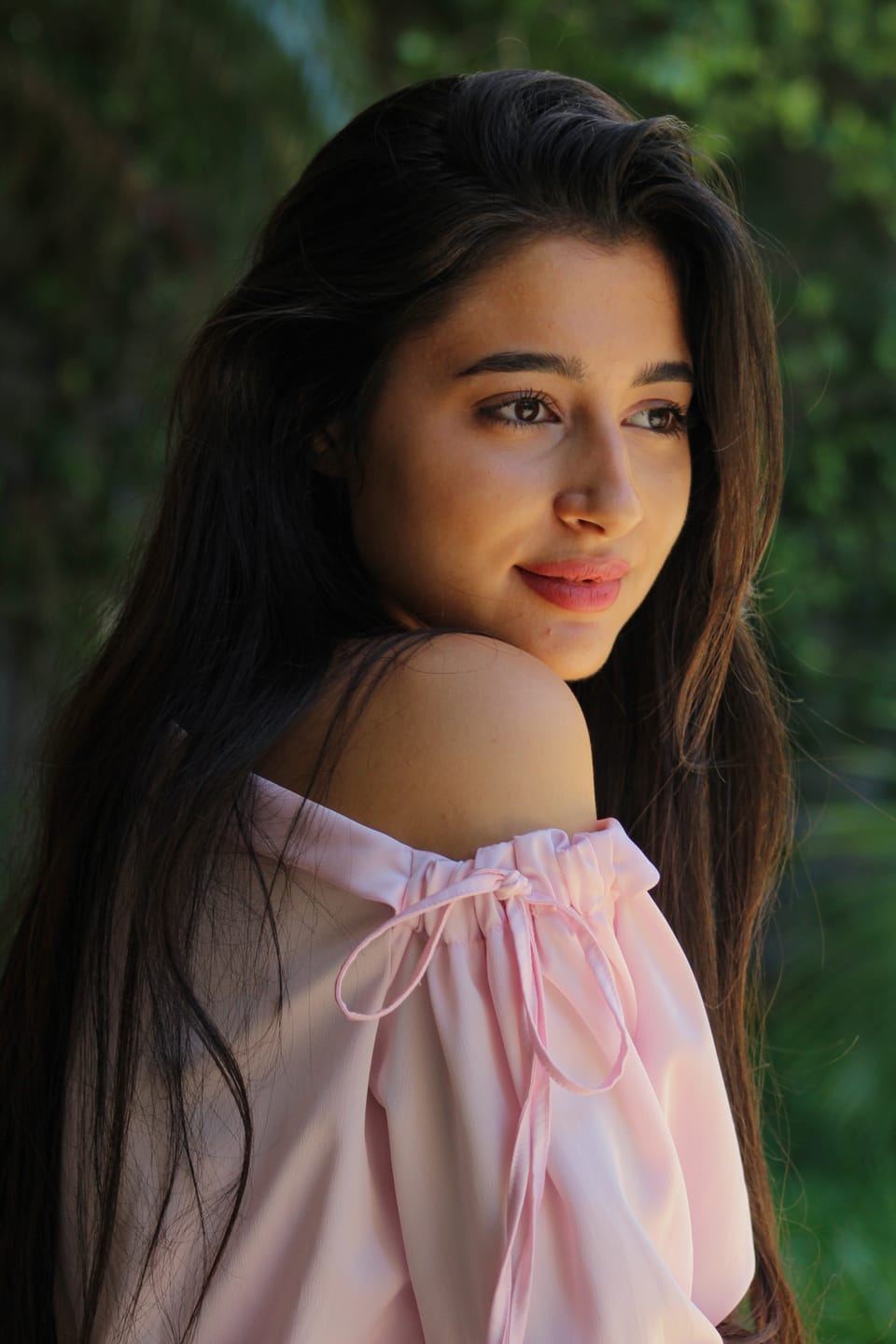 Portrait of a young woman with long dark hair in a pink off-shoulder top, glancing aside against soft green foliage