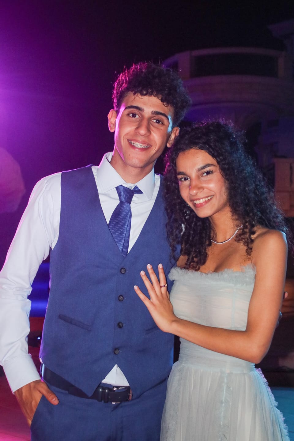 A smiling young couple pose together at a night celebration under purple light