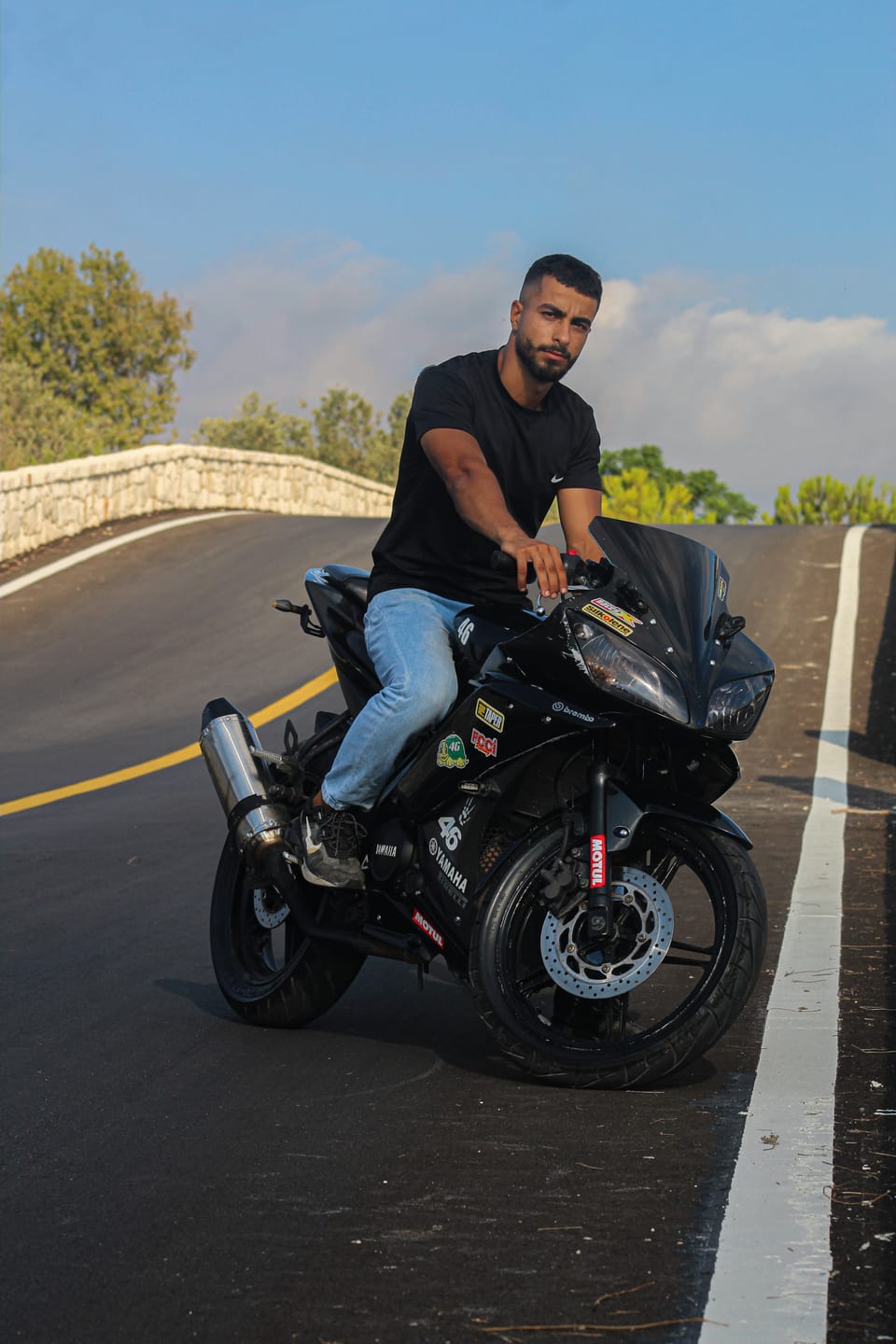 A man in a black t-shirt sits on a black sport motorcycle on an empty curving road under a blue sky