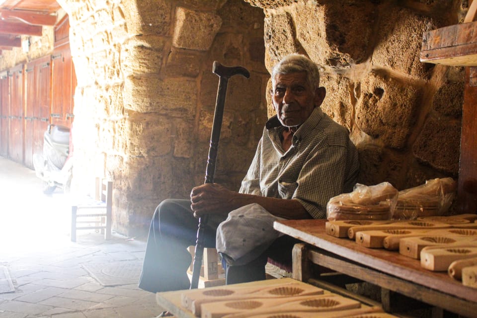 An elderly man with a cane sits beside carved wooden moulds under the stone arch of an old souk