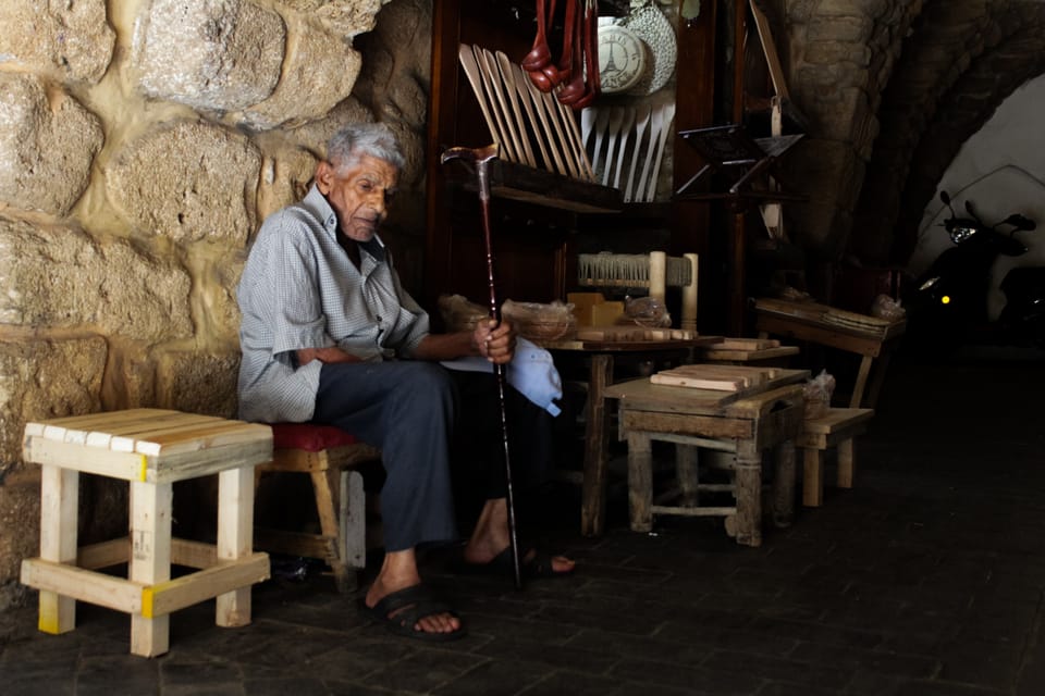 An elderly man rests on a low stool among handmade wooden stools in a dim stone-walled workshop